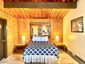 A bedroom with wooden panel walls, a bed with a patterned bedspread, two bedside lamps, a mirror above the bed, and a framed picture on the wall.