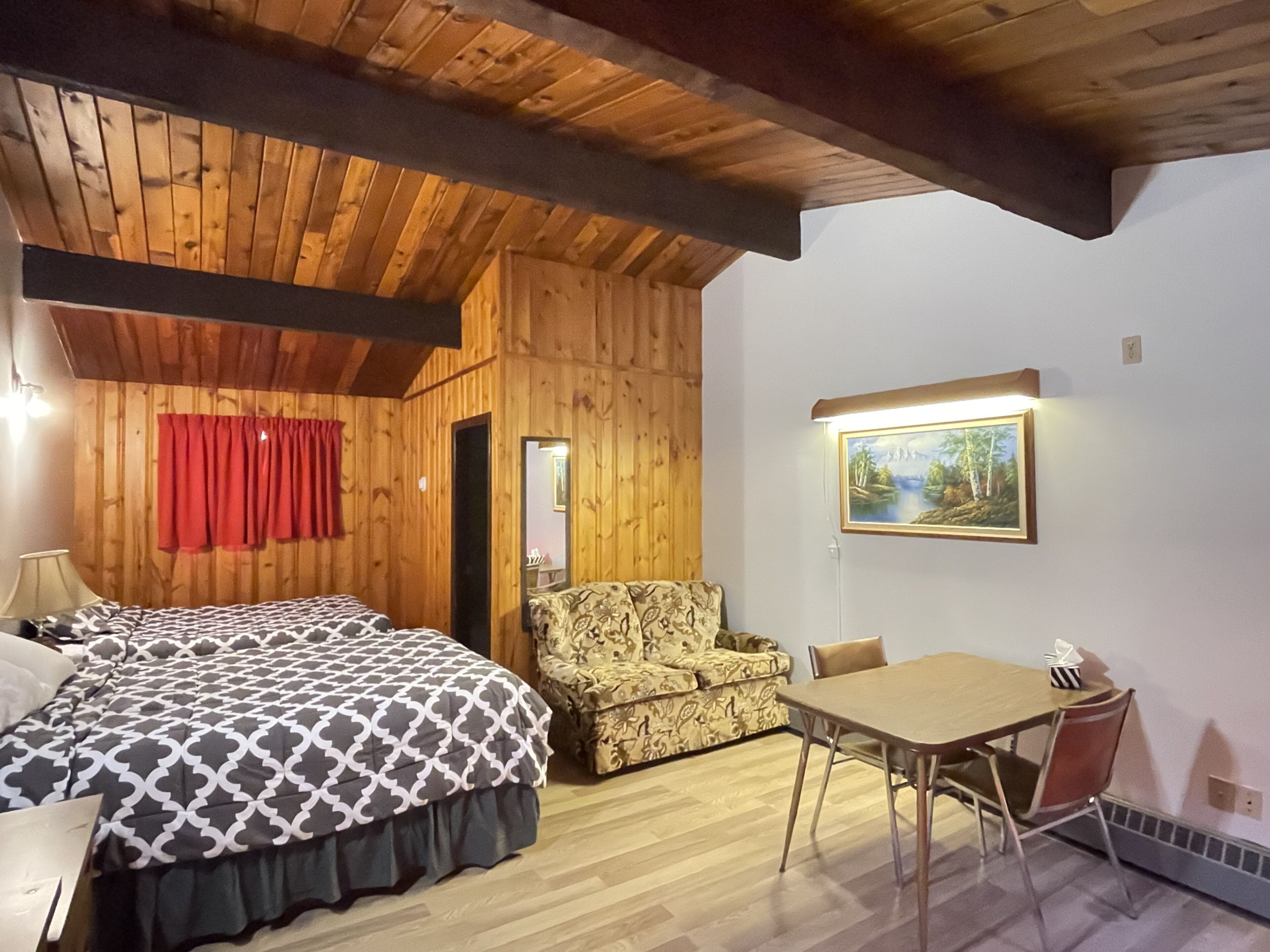 A cozy room with a wooden ceiling, a bed with a patterned bedspread, a sofa, a dining table with chairs, and a painting on the wall.