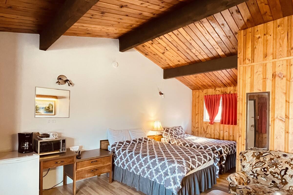 A cozy room with wooden walls and ceiling, a bed with patterned bedding, a desk with a microwave, and a small sofa.