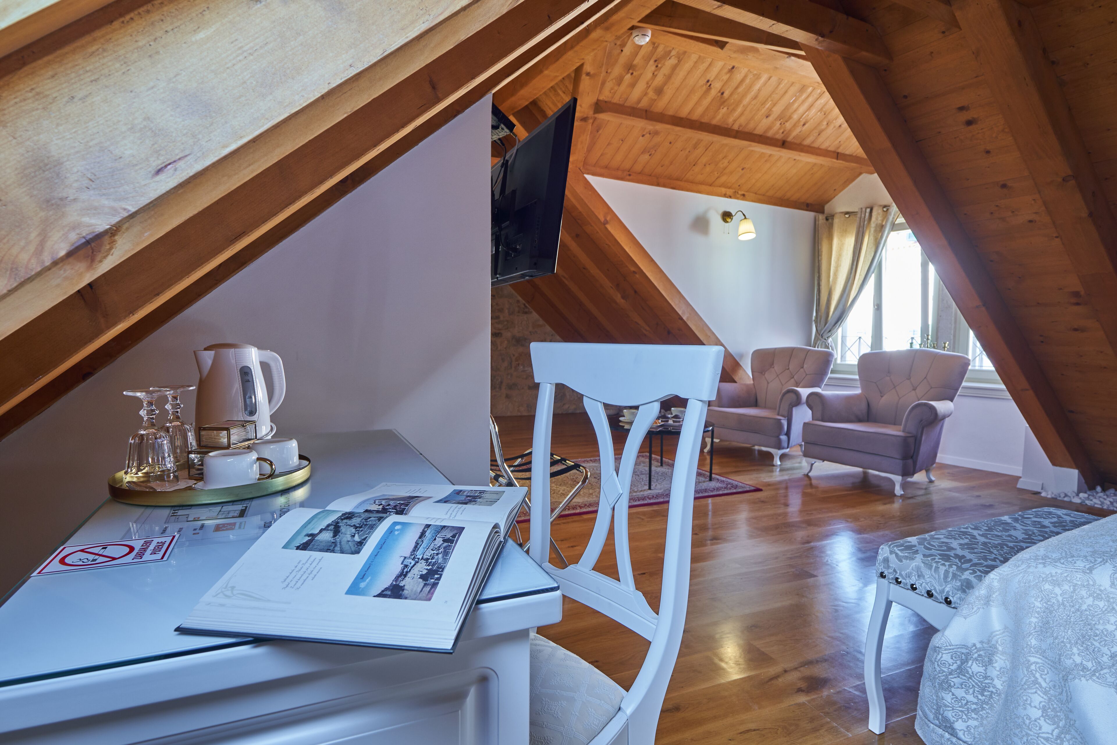 Un acogedor dormitorio en el ático, con vigas de madera, una silla blanca, un sofá y una mesa de comedor con una bandeja de vasos y una tetera.