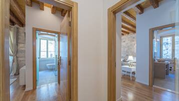A hallway with wooden beams, a glass door leading to a bathroom, and a view of a bedroom with a bed and a nightstand.