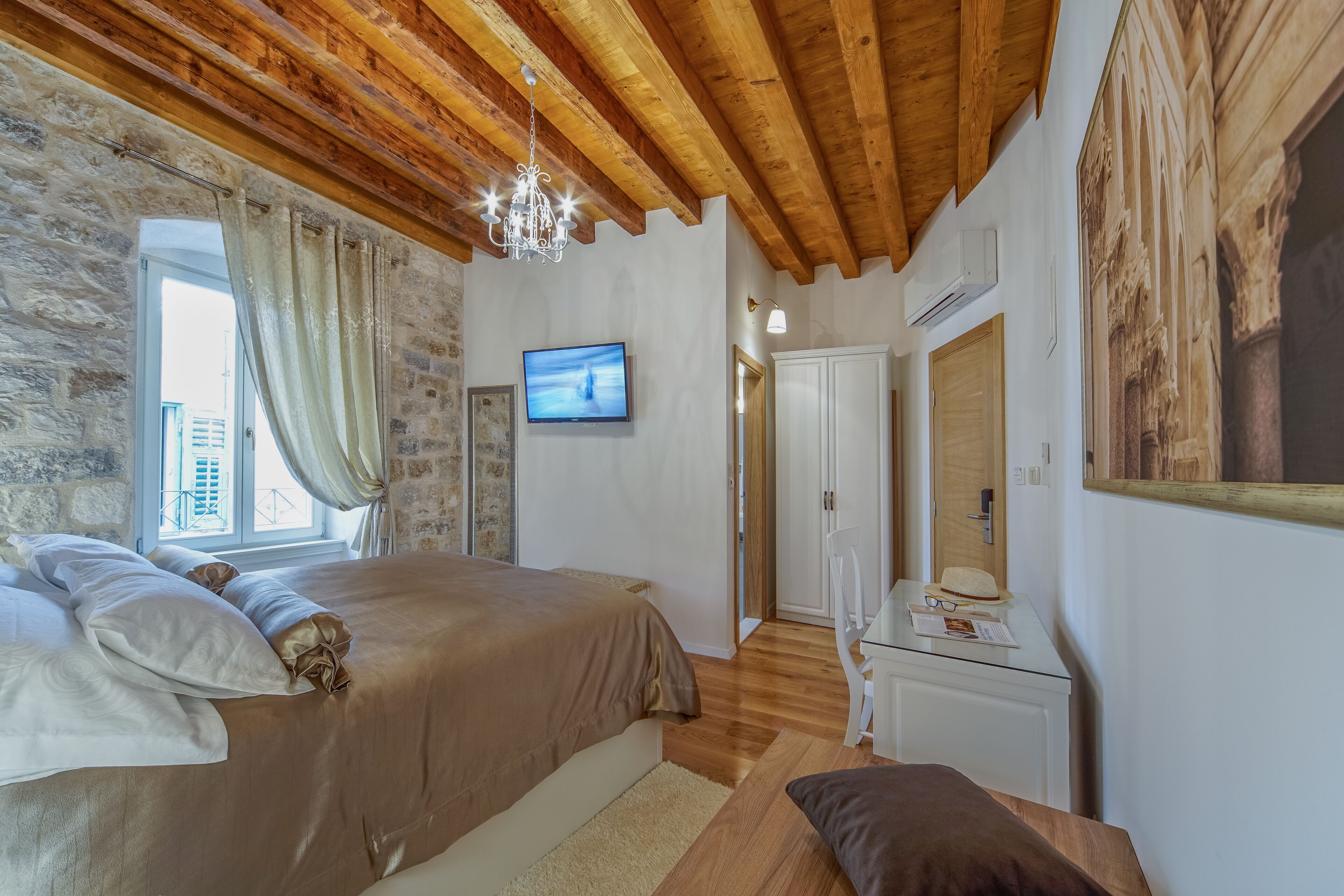 Une chambre avec un plafond en bois, un lit recouvert d’une couverture marron, un téléviseur fixé au mur et une fenêtre avec des rideaux.
