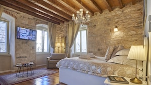 A bedroom with a stone wall, wooden beams, a chandelier, a bed with pillows, a bedside table with a lamp, a TV on the wall, and a small table with cups and bowls. - Villa Split Heritage Hotel (Split)