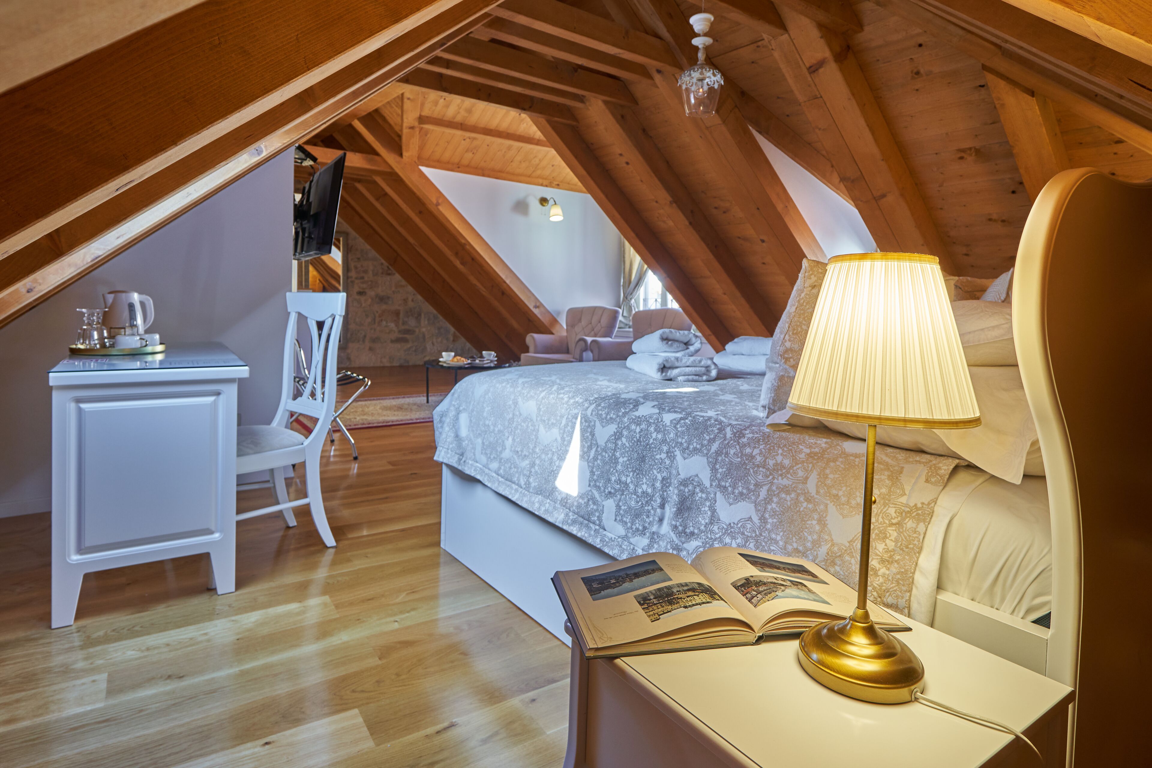 An attic bedroom with a wooden ceiling, a bed with a white headboard, a bedside table with a lamp, and a chair.