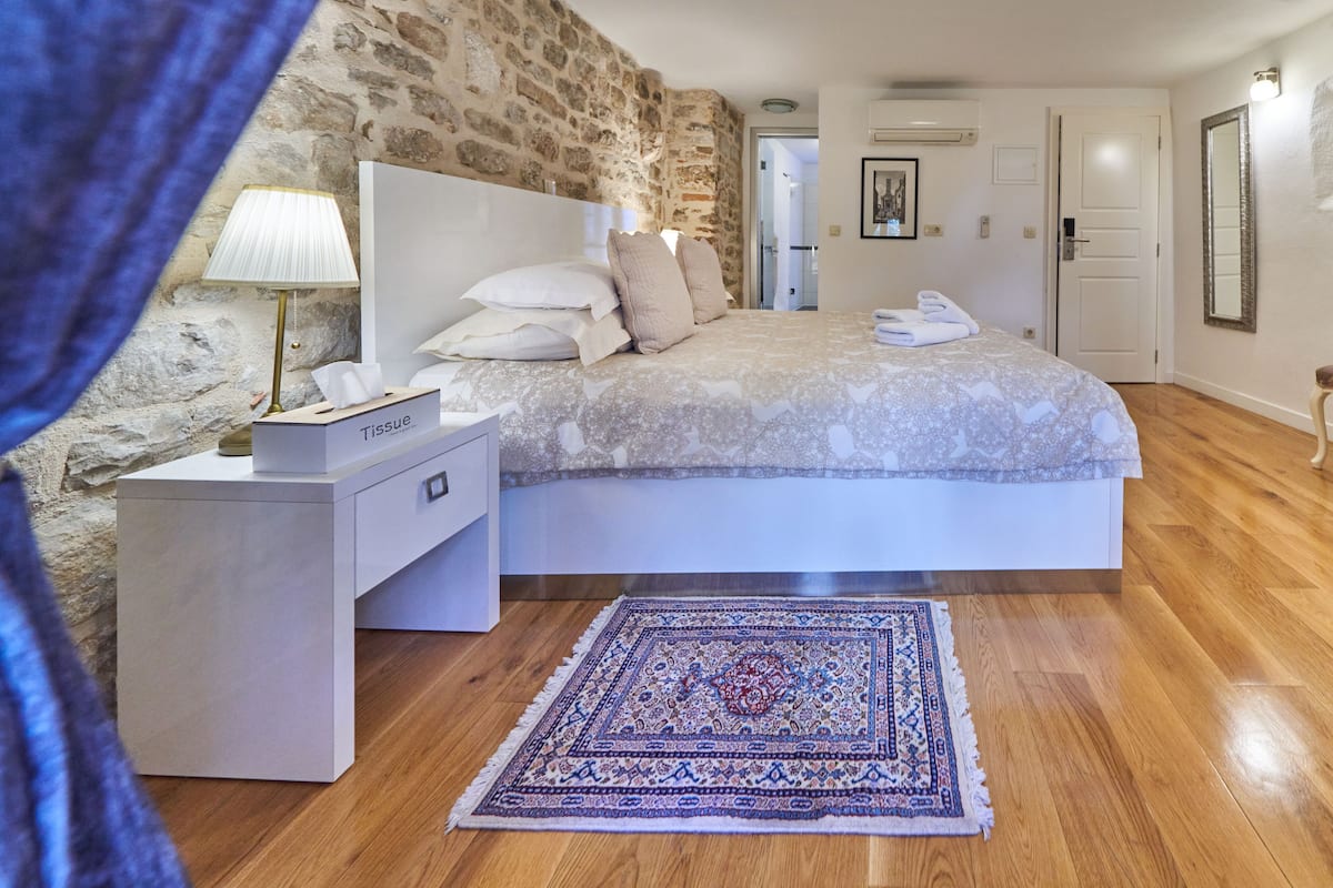 Une chambre avec un mur en pierre, un lit recouvert d’une couvre-lit à motifs, une table de chevet avec une boîte à mouchoirs, un tapis et une porte visible au fond.