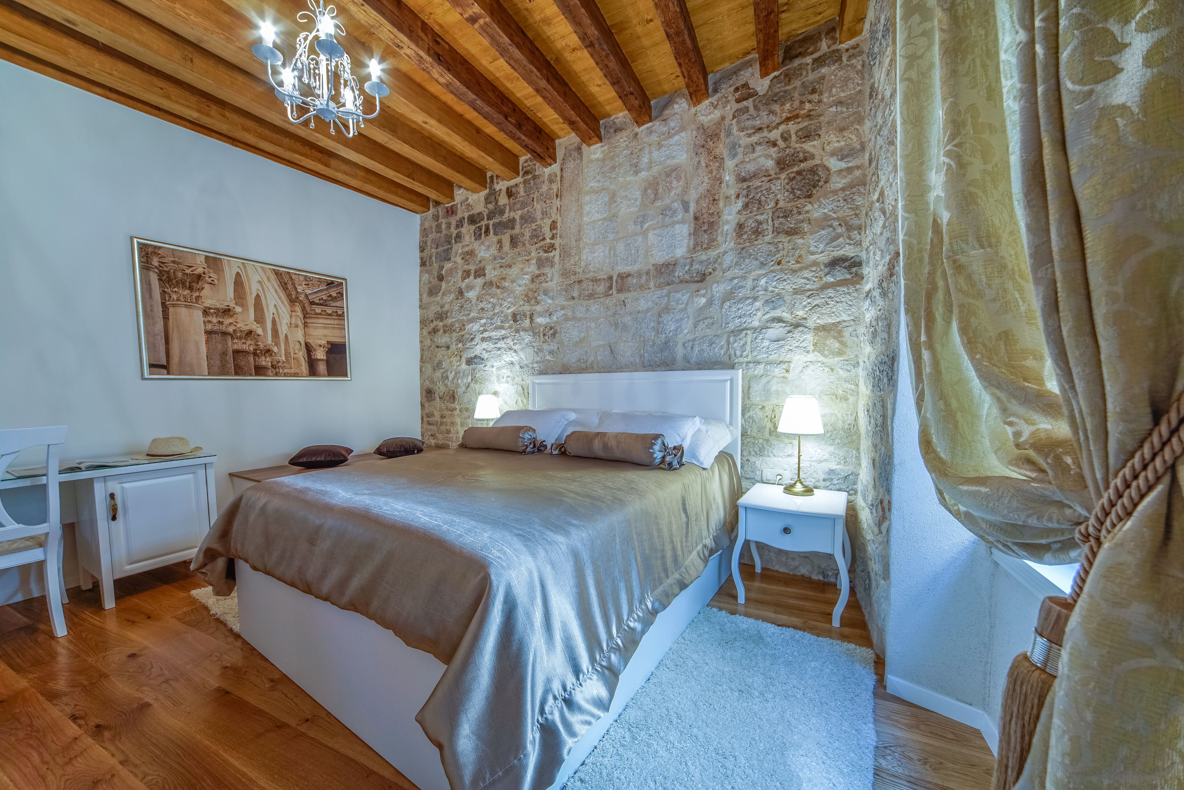 A bedroom with a stone wall, wooden ceiling beams, a bed with a beige bedspread, a desk, and a nightstand with a lamp.