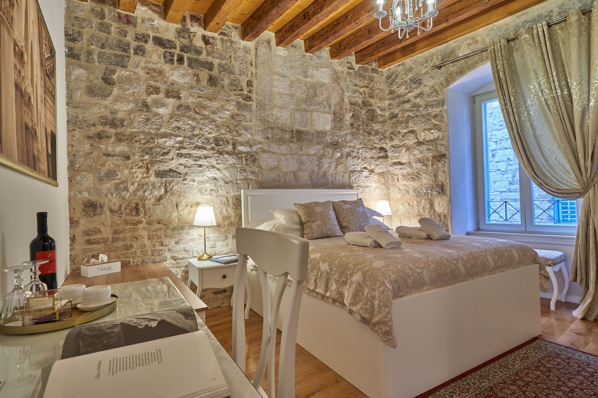 Une chambre avec un mur en pierre, un plafond en bois, un lit recouvert d’une couvre-lit beige, une table de chevet avec une lampe et une fenêtre avec des rideaux.