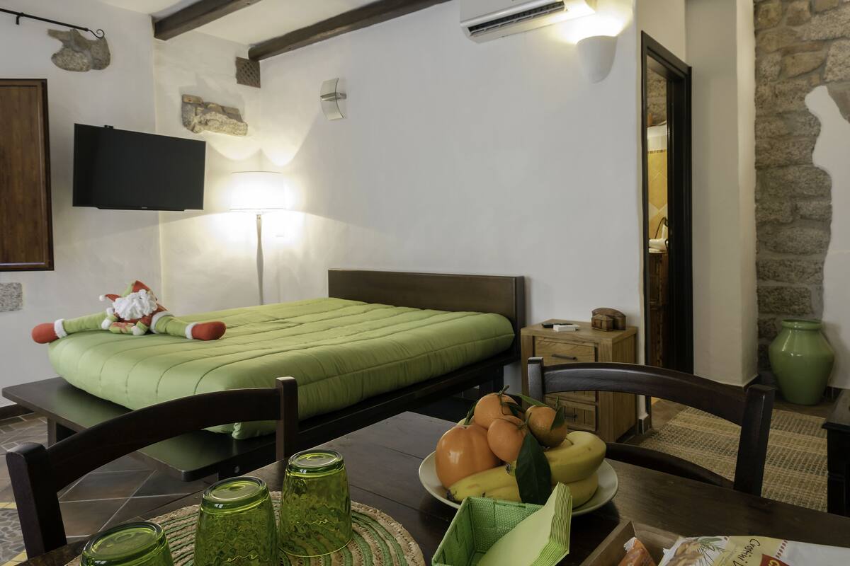 A bedroom with a green bed, a flat-screen TV, a stone wall, and a dining area with a table set with fruits and a green vase.