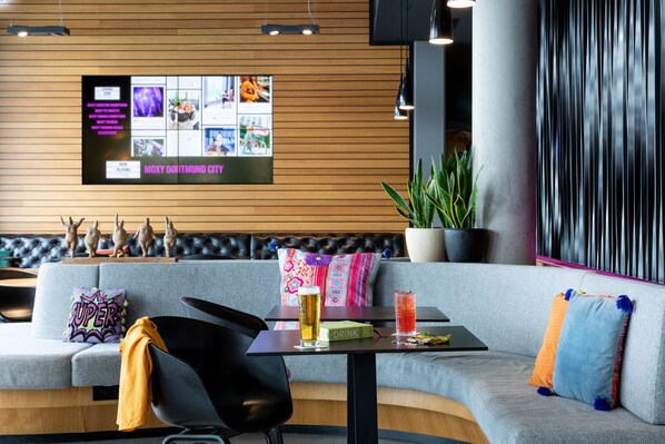 A modern lounge area with a grey sofa, black chairs, a table with a drink and a menu, and a wall-mounted screen displaying images and text.