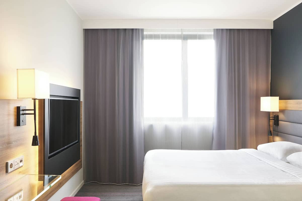 A hotel room with a bed, a TV mounted on the wall, a window with curtains, and a lamp on the bedside table.