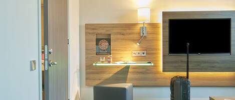A modern hotel room with a wooden accent wall, a flat-screen TV, a bedside table with a lamp, a small ottoman, and a suitcase.