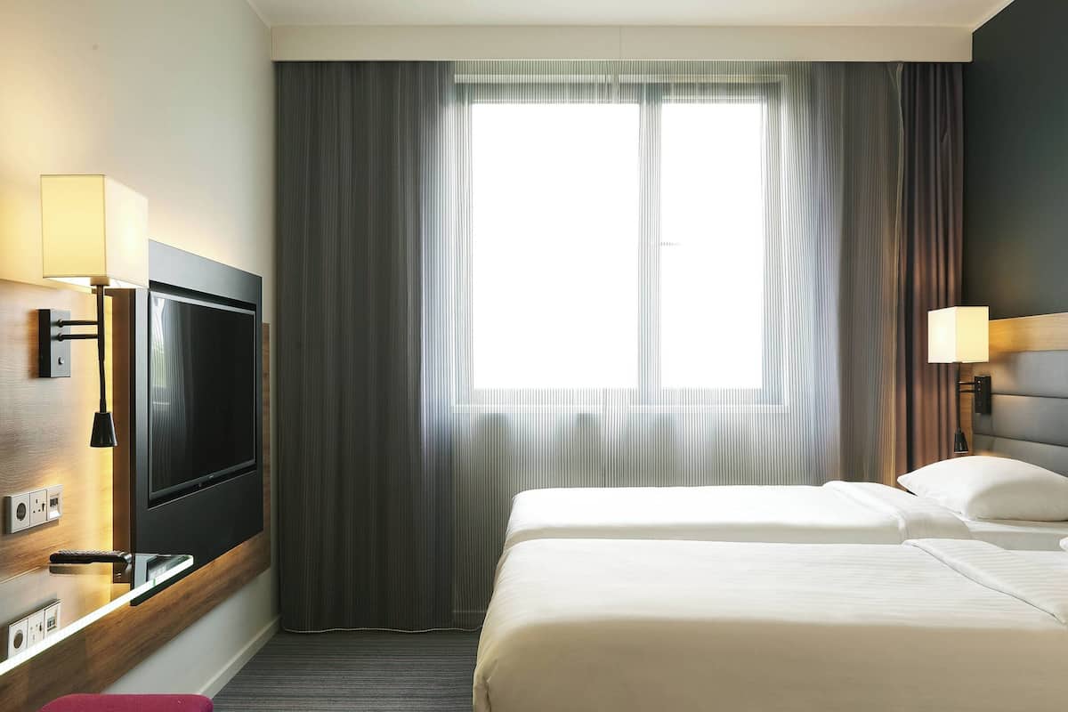 A hotel room with a TV mounted on the wall, a bed with white bedding, and a window with curtains.