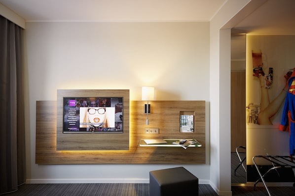 Habitación de hotel con televisor montado, lámpara de noche, revistero y perchero con una chaqueta.