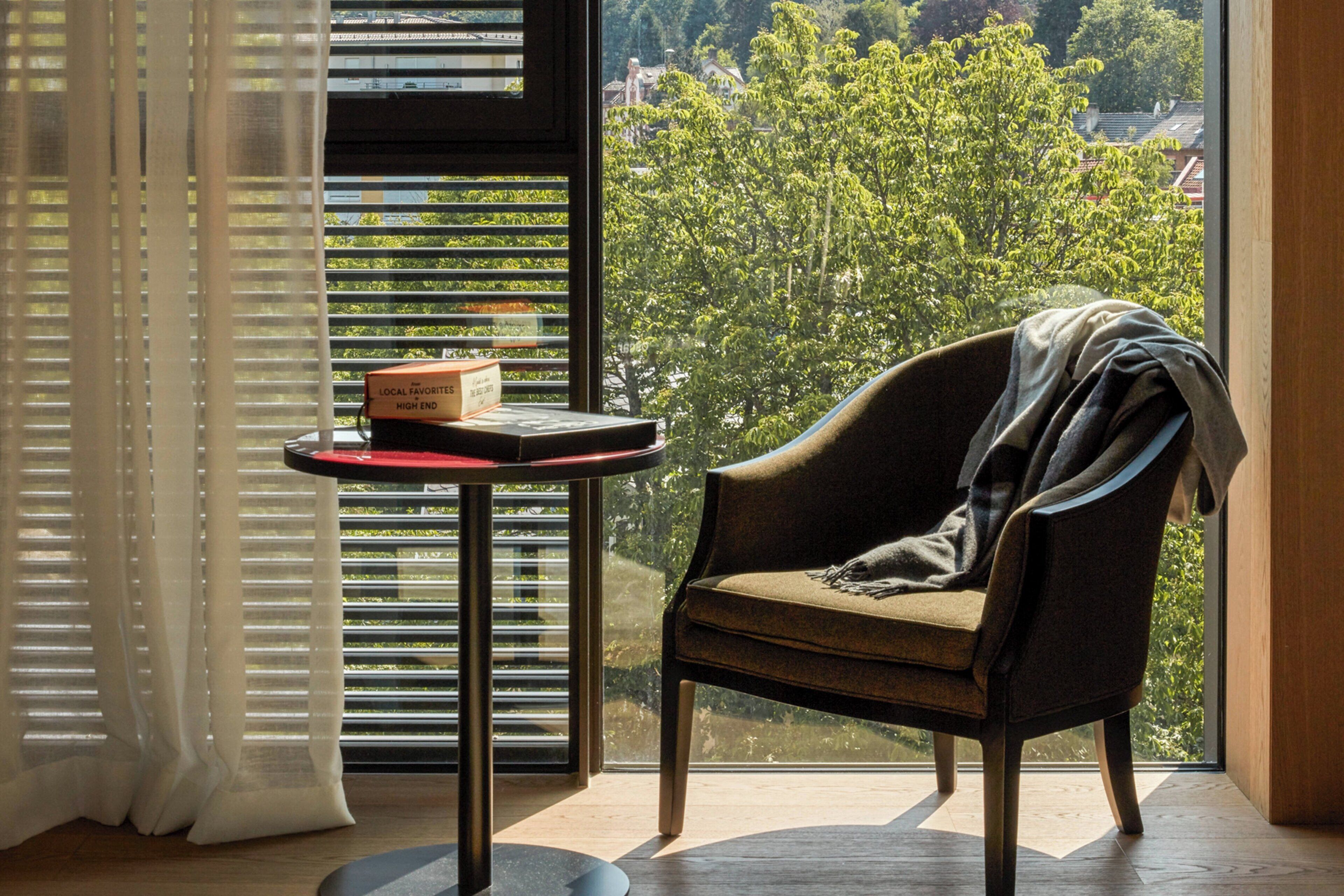Una habitación con vistas a árboles y edificios, una silla, una mesa con una caja y una ventana con persianas.