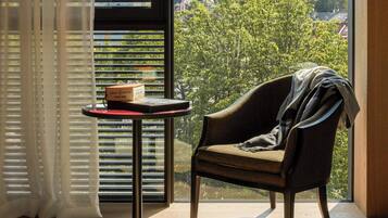 Una habitación con vistas a árboles y edificios, una silla, una mesa con una caja y una ventana con persianas.