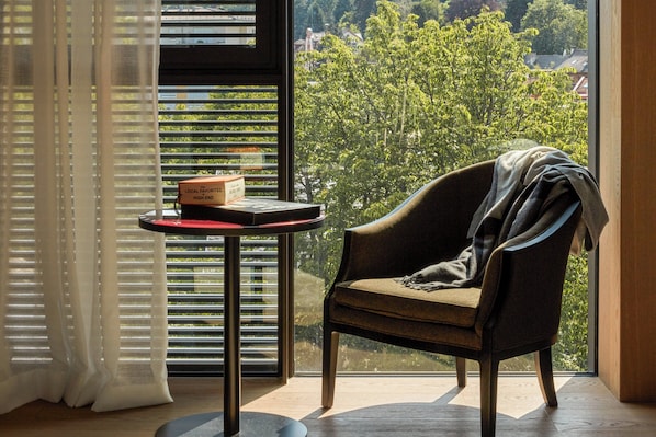 Una habitación con vistas a árboles y edificios, una silla, una mesa con una caja y una ventana con persianas.