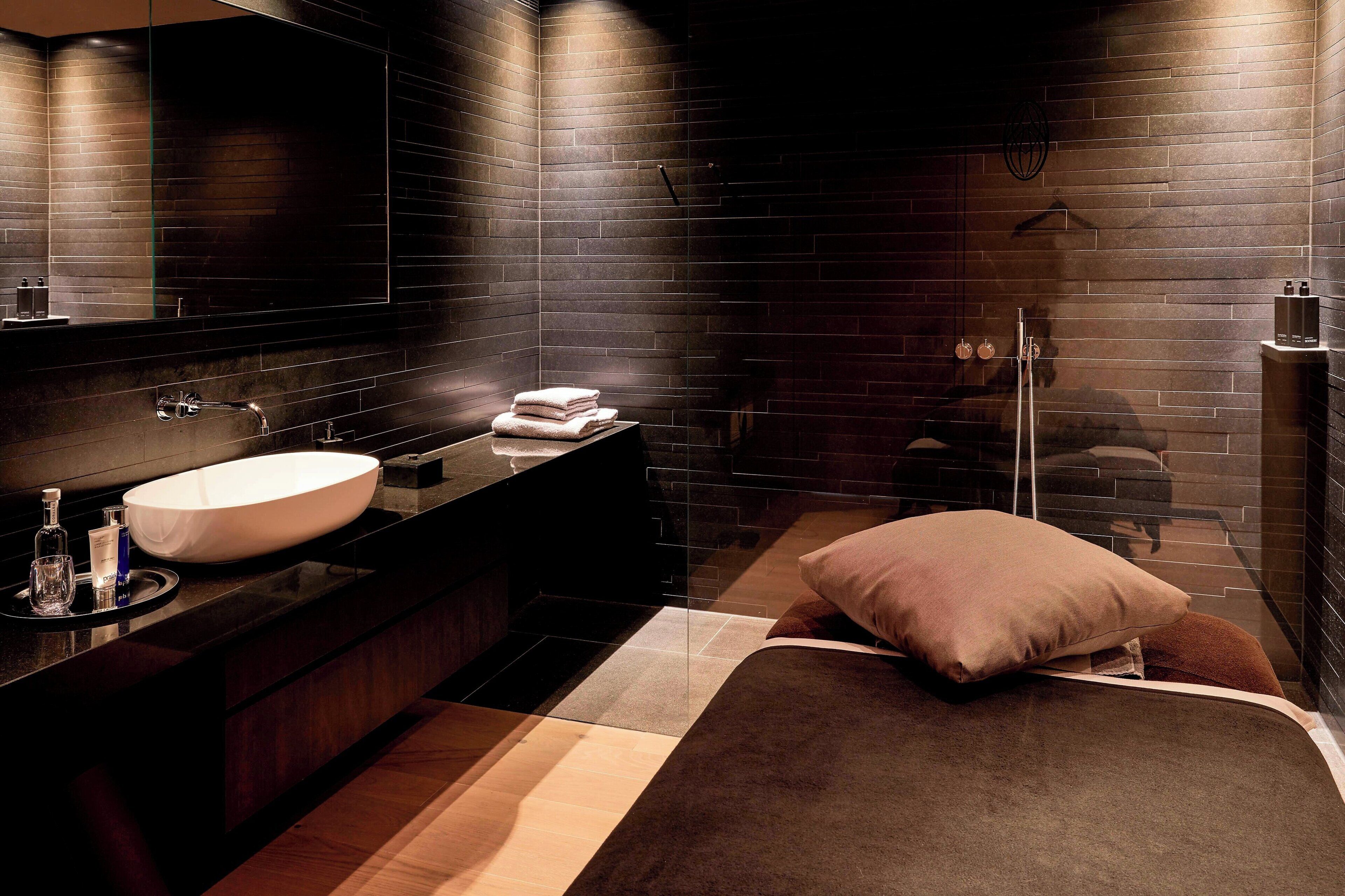 A modern bathroom with a large mirror, dark brick walls, a white sink, and a wooden floor.