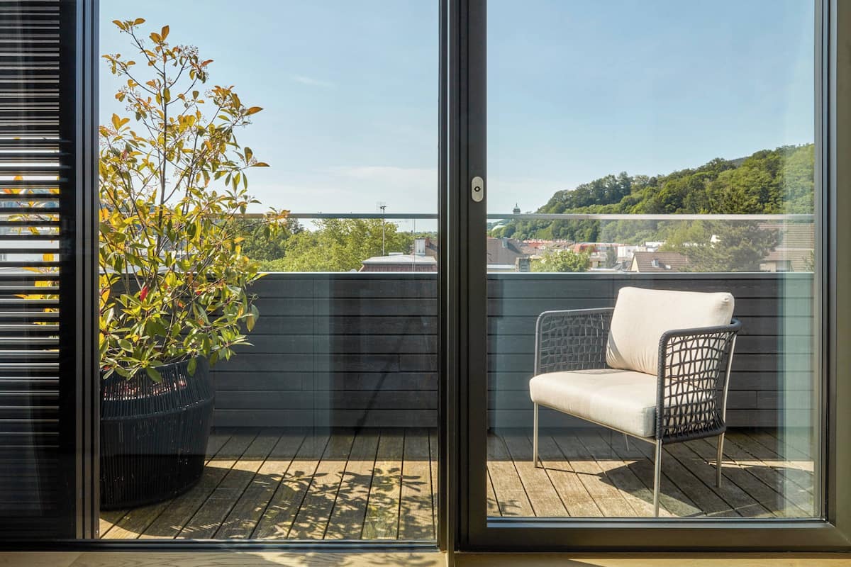 Un balcon agrémenté d’un pot de fleurs et d’une chaise, d’où l’on peut admirer un paysage urbain avec ses bâtiments et sa verdure.