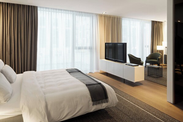Una habitación de hotel moderna con una cama grande, un televisor de pantalla plana montado en un mueble blanco, dos sillones y un escritorio con una lámpara.