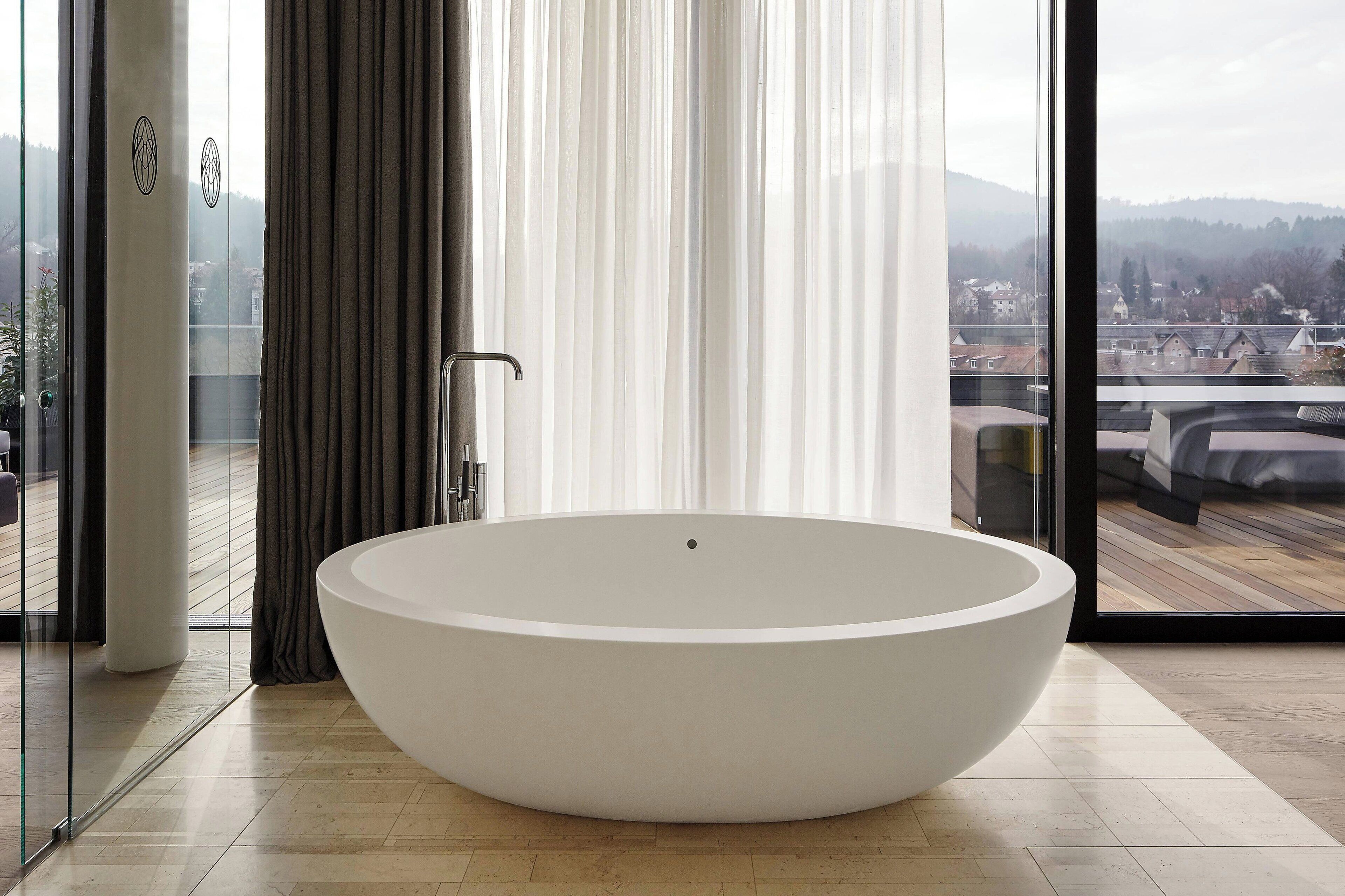 Un baño moderno con una bañera ovalada grande, un box de ducha de vidrio y vistas a una zona residencial a través de amplios ventanales.
