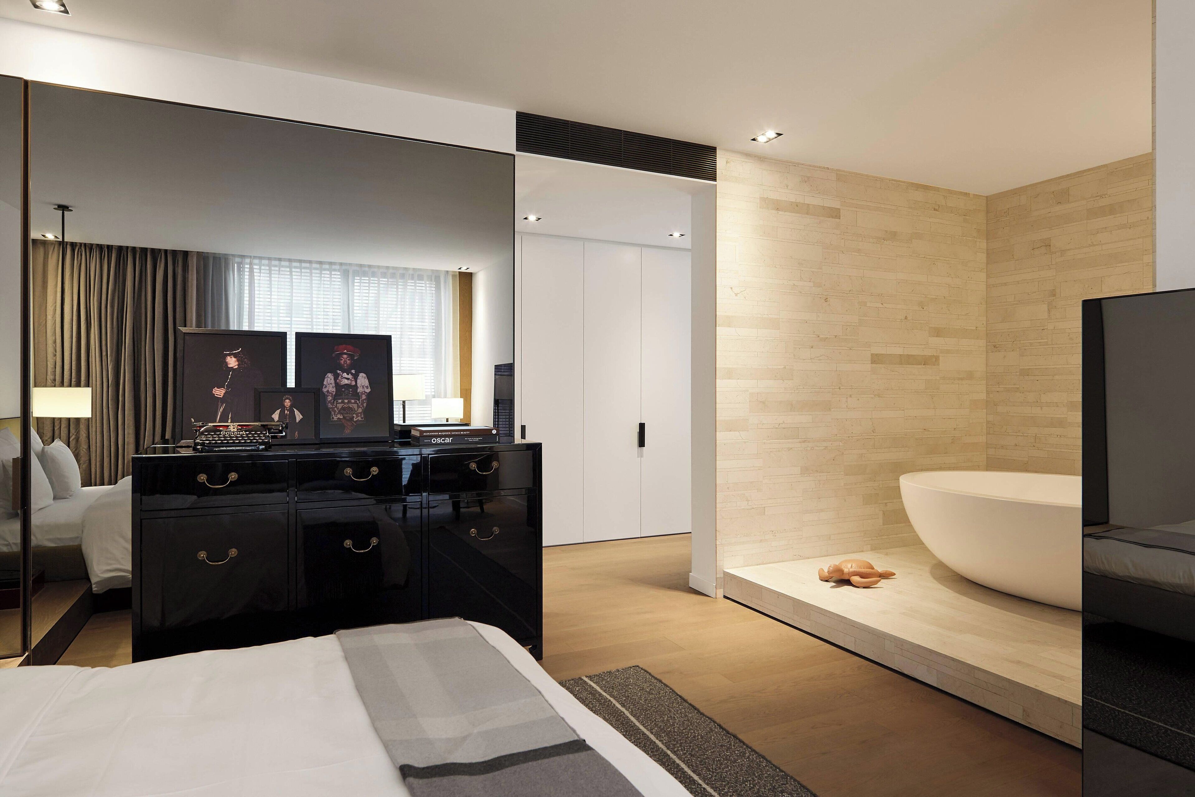 Une chambre d’hôtel moderne avec un grand lit, une commode noire, un grand miroir, une armoire blanche, un mur d’accent en bois et une grande baignoire.