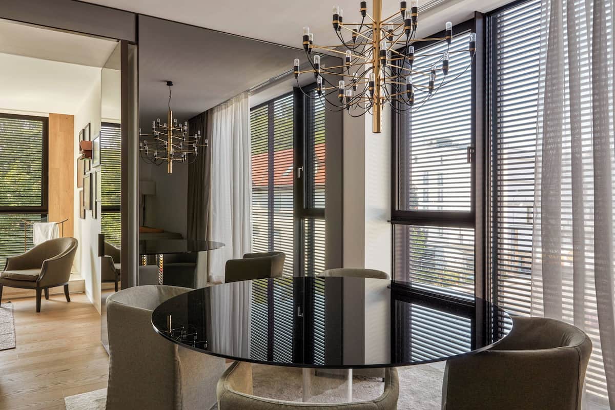 Un espace de salle à manger moderne avec une table en verre entourée de chaises, de grandes fenêtres équipées de stores et un lustre.