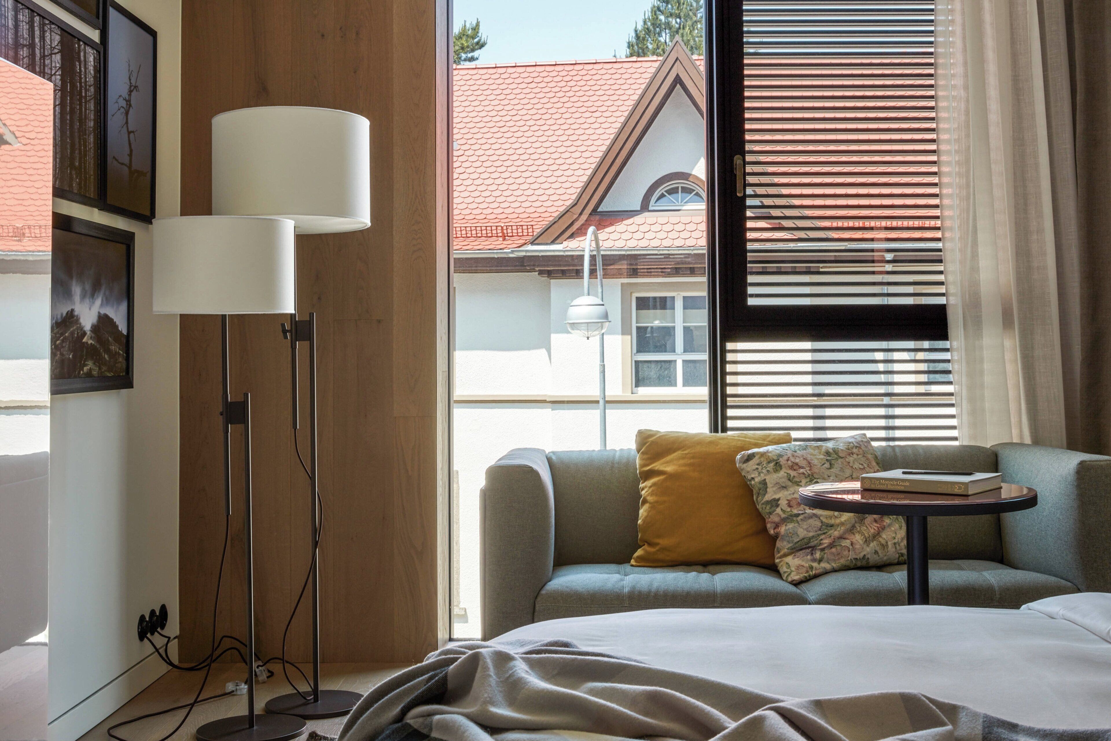 Een hotelkamer met een bank, een kleintje tafeltje, een lamp en een raam met zonwering.