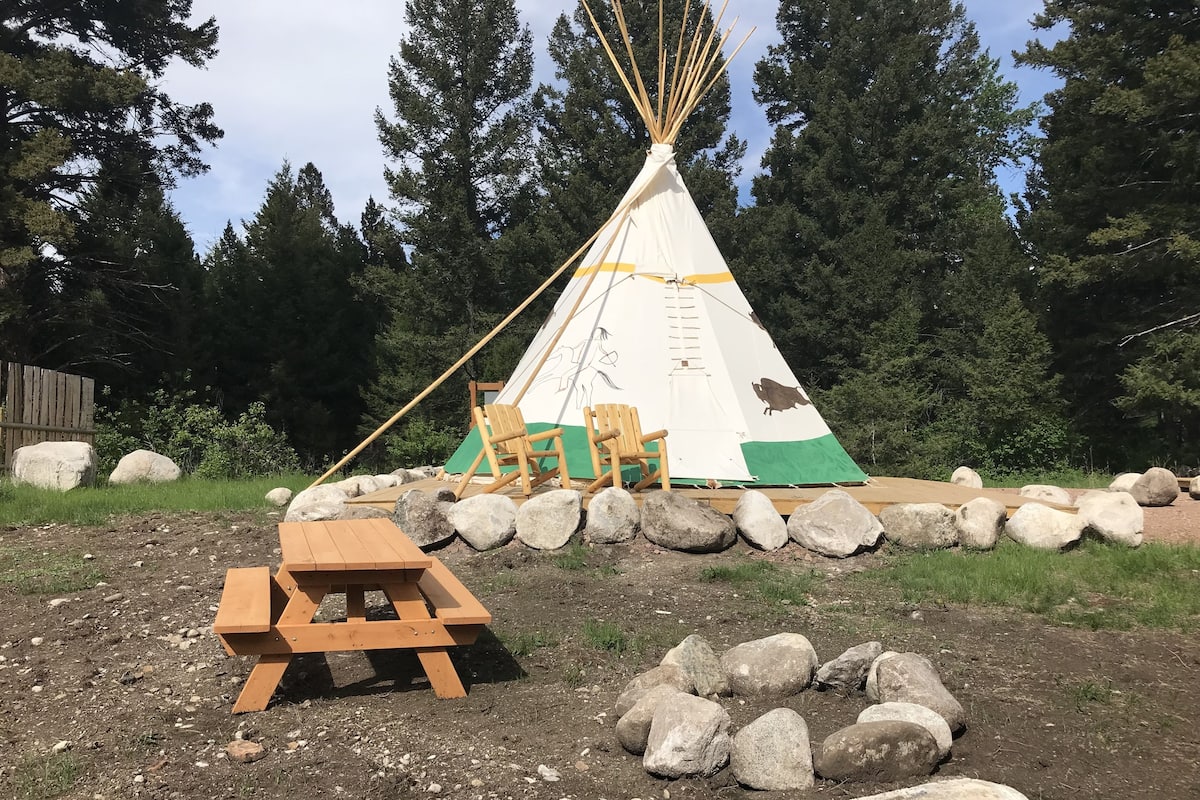 Uma tenda tipi com uma mesa de piquenique e duas cadeiras na frente, cercada por pedras e árvores.