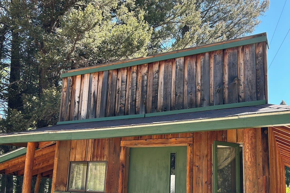 Uma cabana de madeira rústica com porta verde, um pequeno alpendre com cadeira de madeira e uma placa com a inscrição "The Creek".