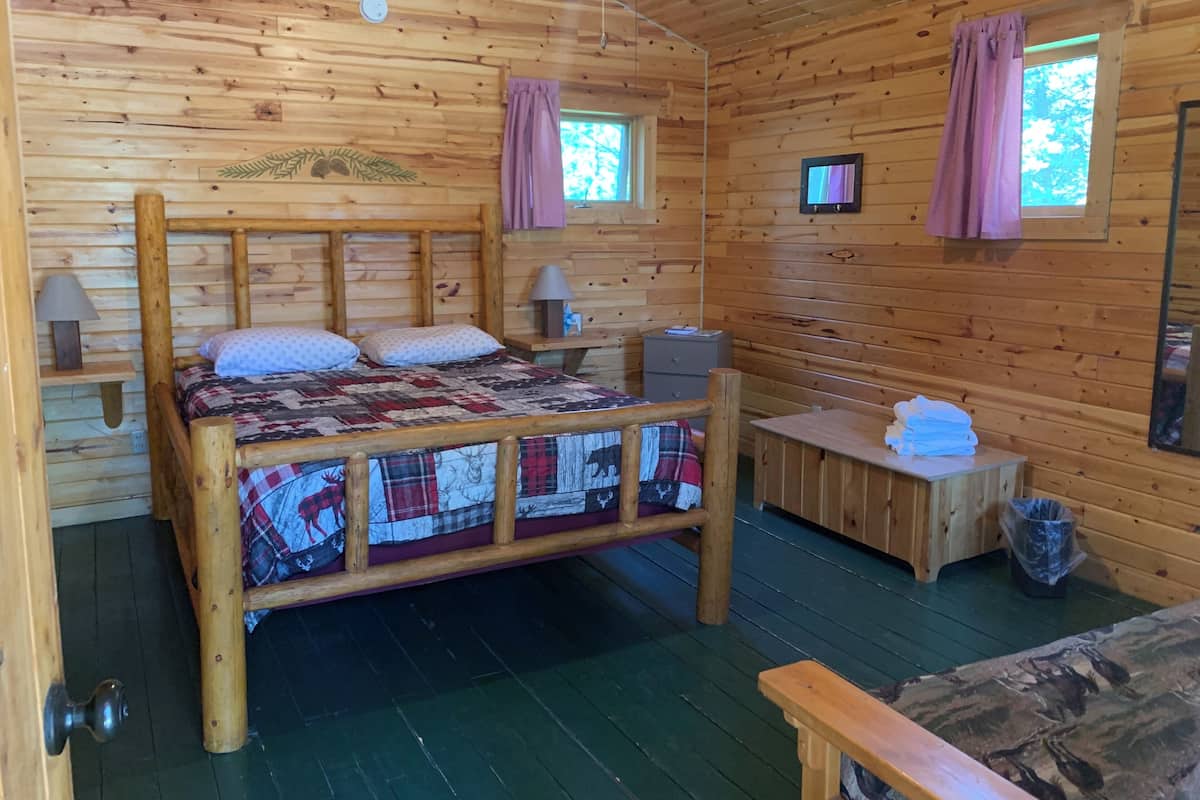 Quarto de cabana de madeira com cama, luminárias de cabeceira, uma mesinha e uma janela com cortinas.