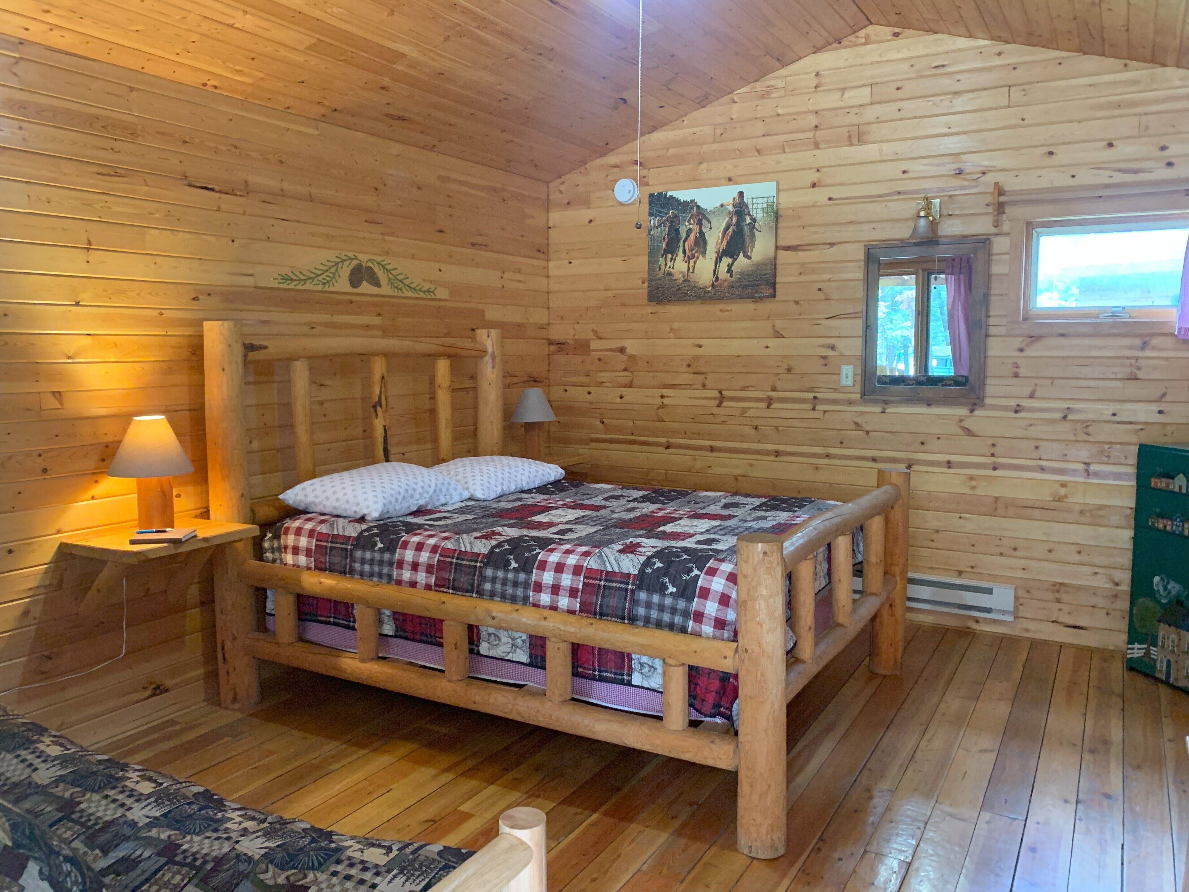 Ein Schlafzimmer einer Holzhütte mit einem Bett, einem Nachttisch, einer Lampe, einem Fenster und einem Gemälde an der Wand.