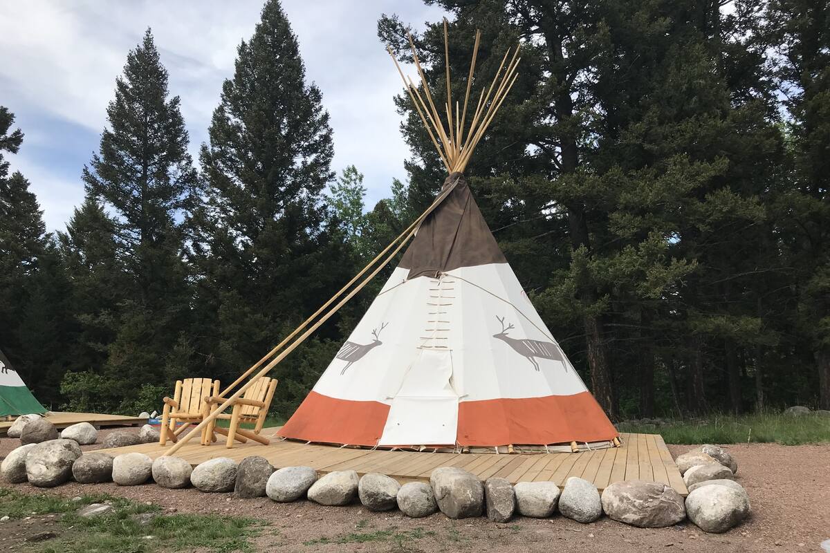 Uma tenda tipi com desenho de veado, cercada por pedras e um deck de madeira.