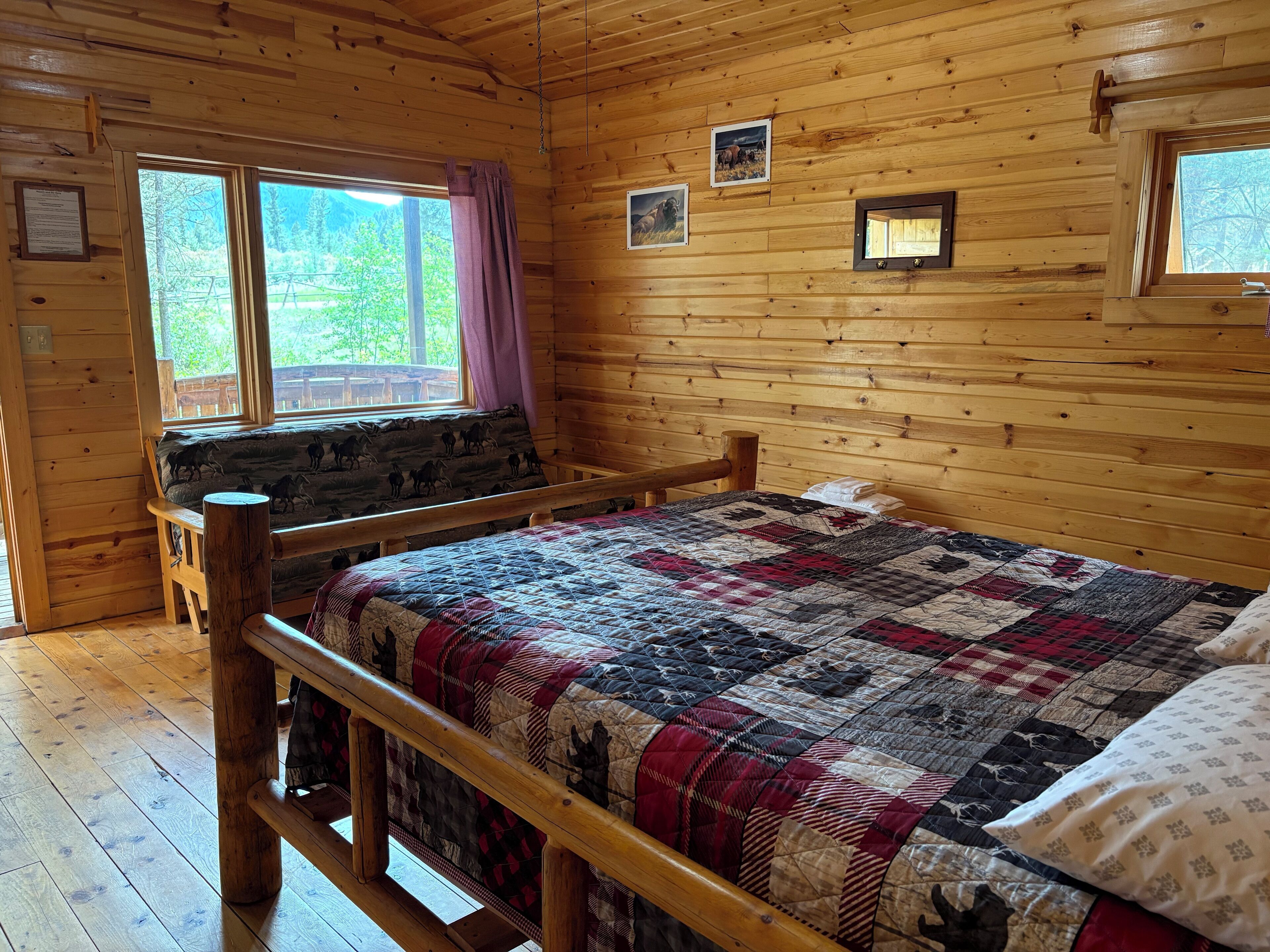 A wooden cabin bedroom with a bed, a bench, and a window with a view of greenery.