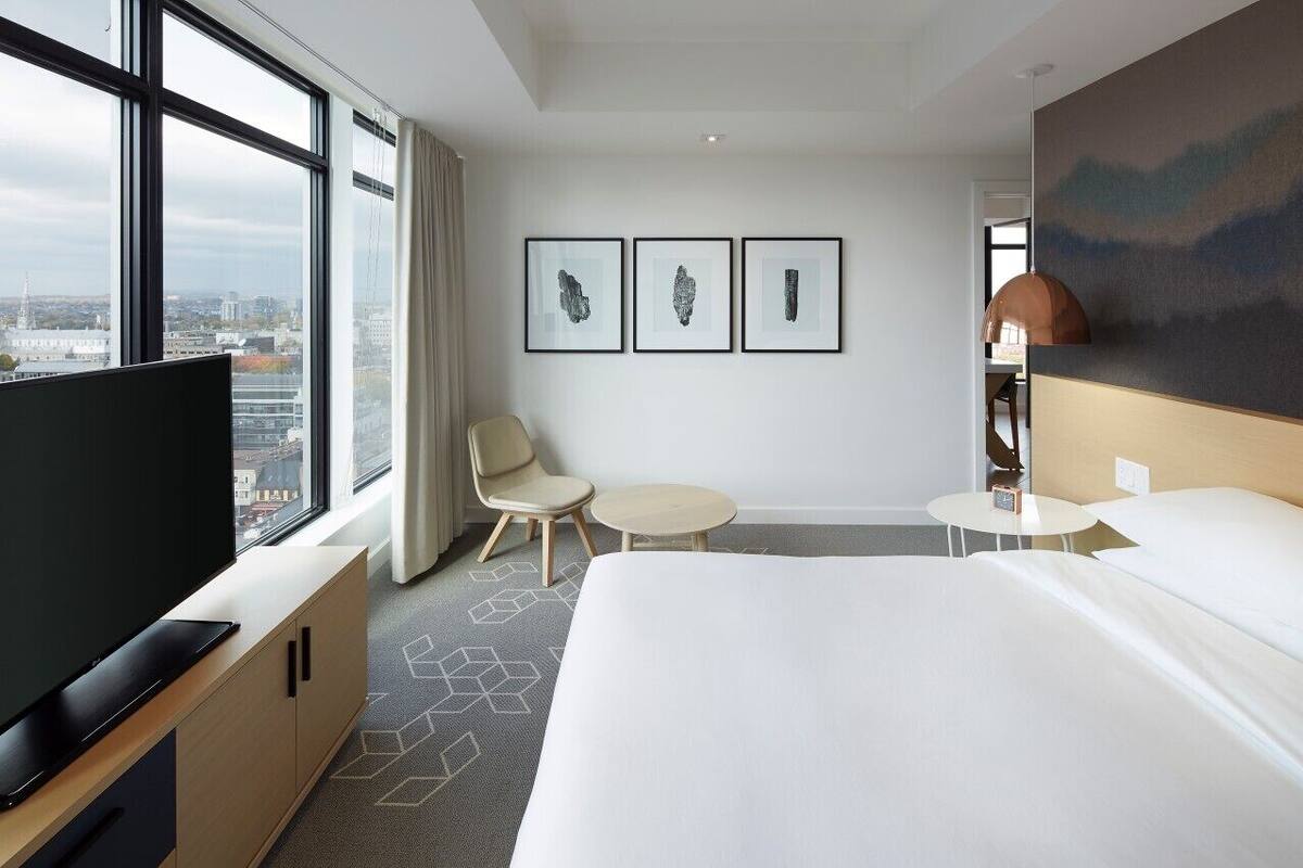 A hotel room with a large bed, a TV on a wooden cabinet, a desk, two chairs, and a city view through large windows.