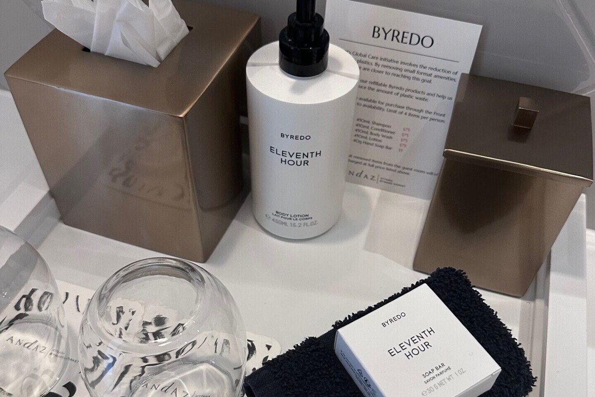 Baño de una habitación de hotel con artículos de aseo que incluyen una botella de loción corporal BYREDO ELEVENTH HOUR, una pastilla de jabón, una toalla y una caja de pañuelos.
