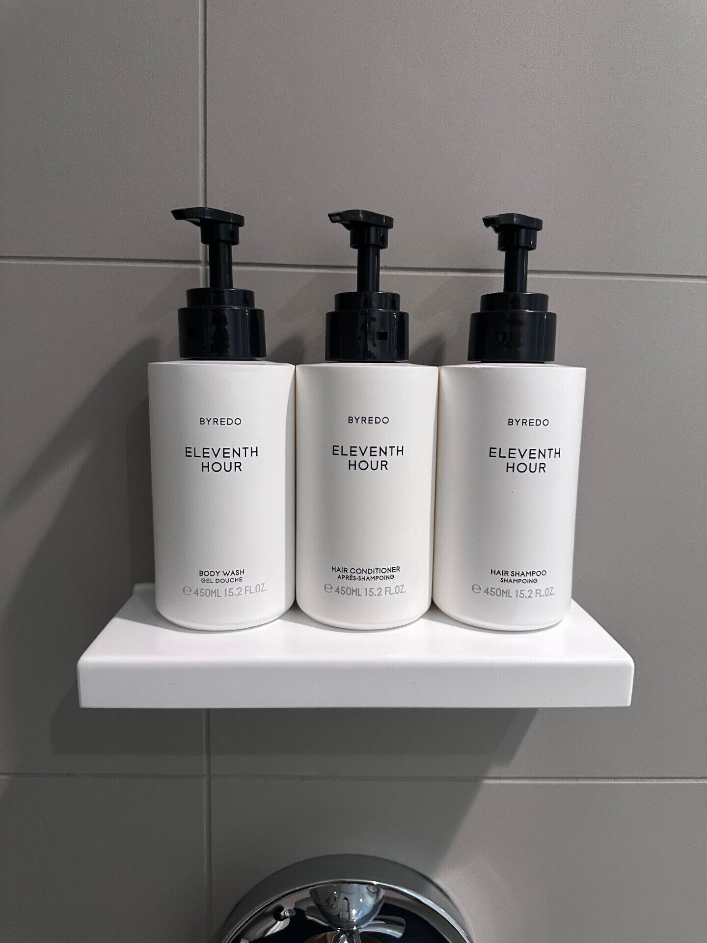Três garrafas brancas de BYREDO Eleventh Hour numa prateleira branca, em frente a uma parede revestida de azulejos.