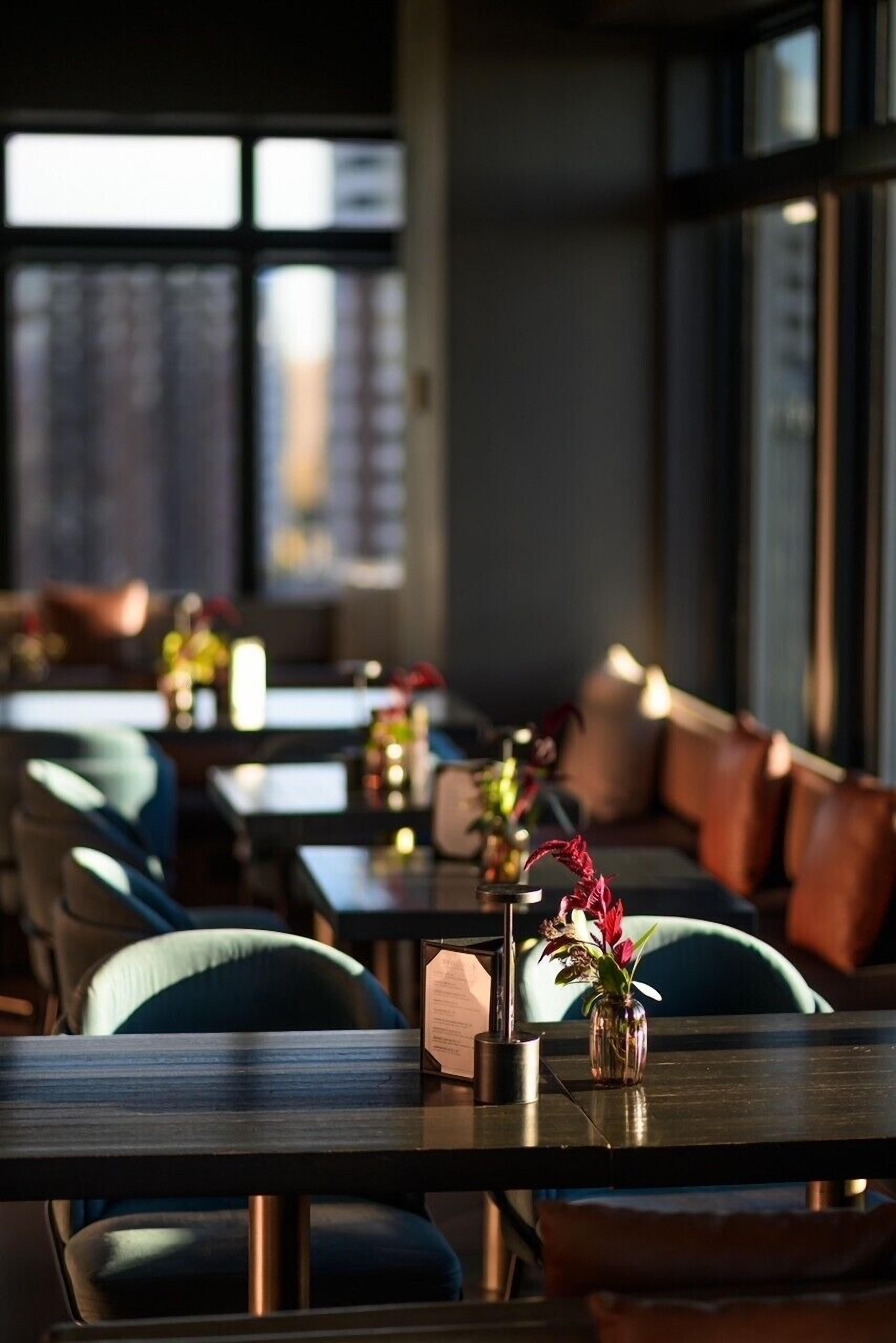 Un restaurante moderno con mesas largas, asientos acolchados y un jarrón con flores en cada mesa.