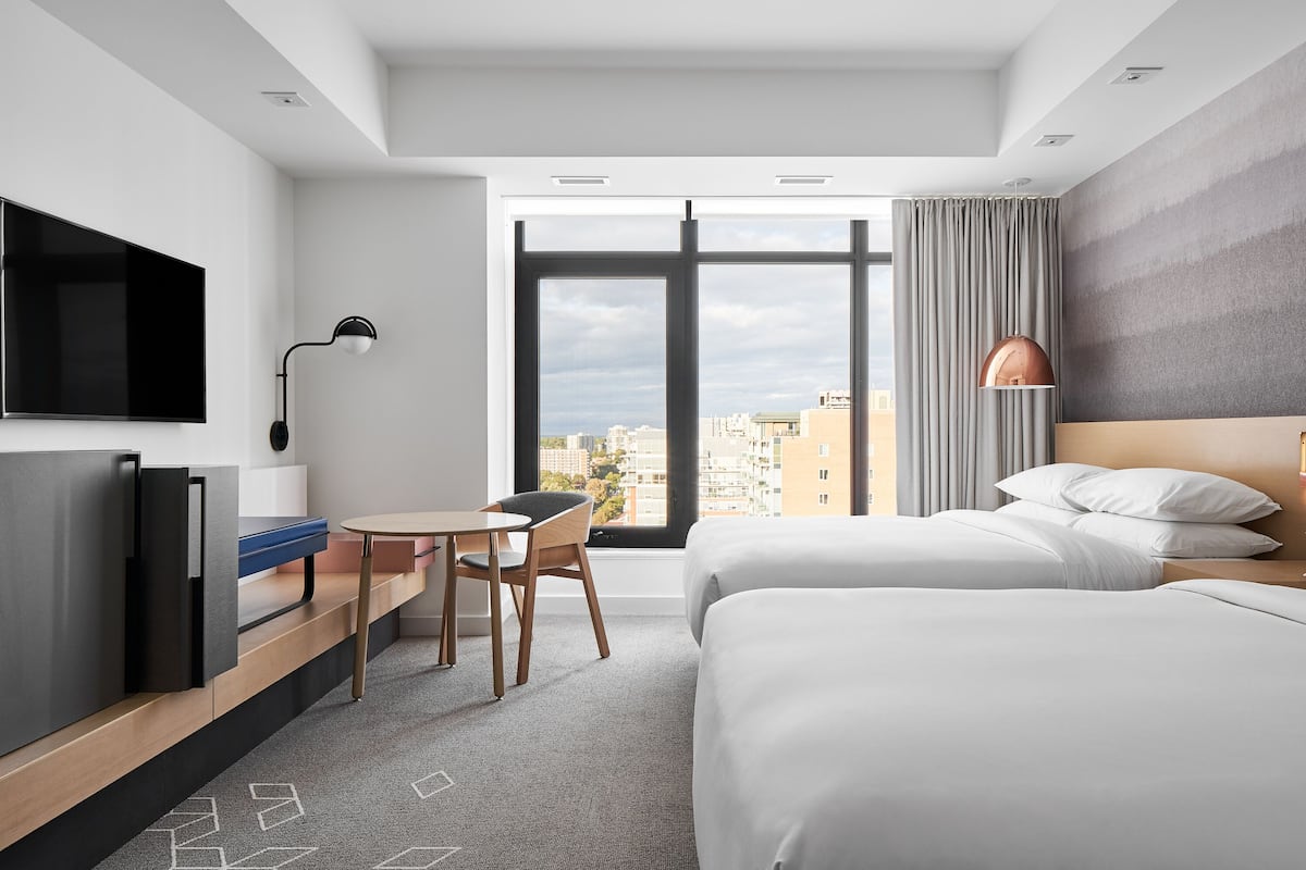 A modern hotel room with a flat-screen TV, a desk with a chair, two beds, a large window with curtains, and a city view.