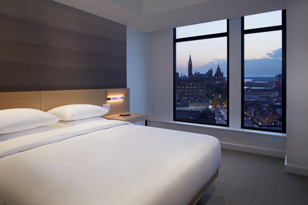 A hotel room with a large bed, a nightstand, a lamp, and a window with a city view.