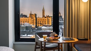 Habitación de hotel con un amplio ventanal que ofrece vistas a un edificio histórico. Hay una mesa de comedor con alimentos para el desayuno y una computadora portátil.