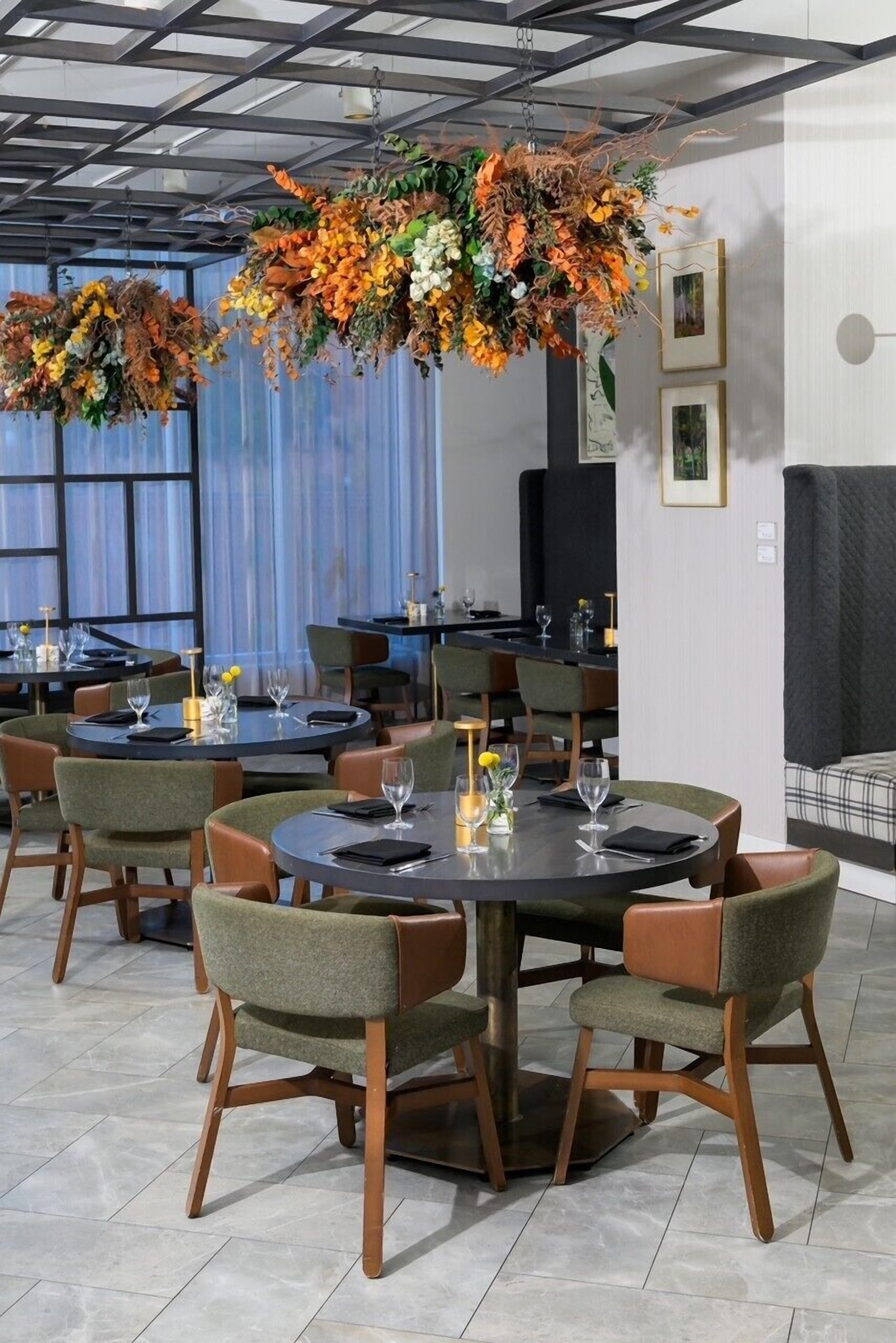 A restaurant with round tables, green upholstered chairs, and hanging floral arrangements.