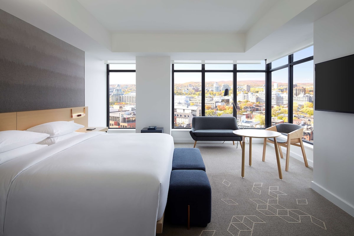 A modern hotel room with a large bed, a small table, two chairs, and a view of the city through large windows.