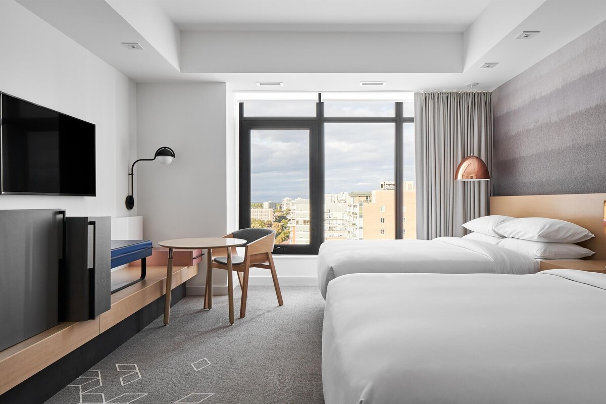 A modern hotel room with a bed, a desk, a television, and a large window offering a city view.