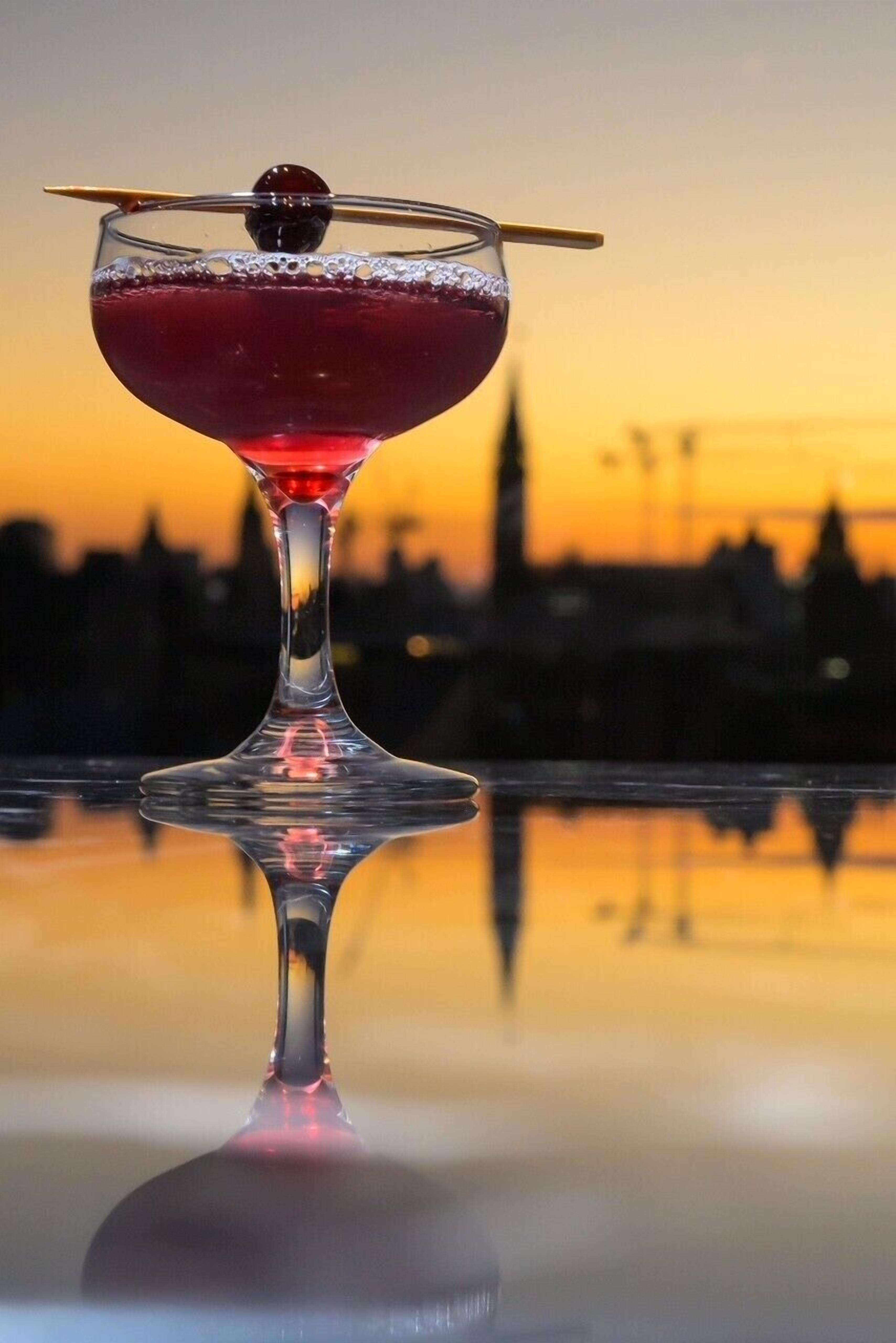 A cocktail in a stemmed glass with a cherry garnish, set against a sunset skyline.