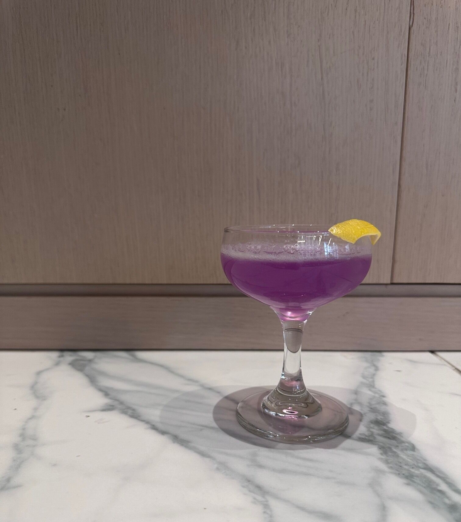 A purple drink in a stemmed glass with a lemon garnish on a marble countertop.