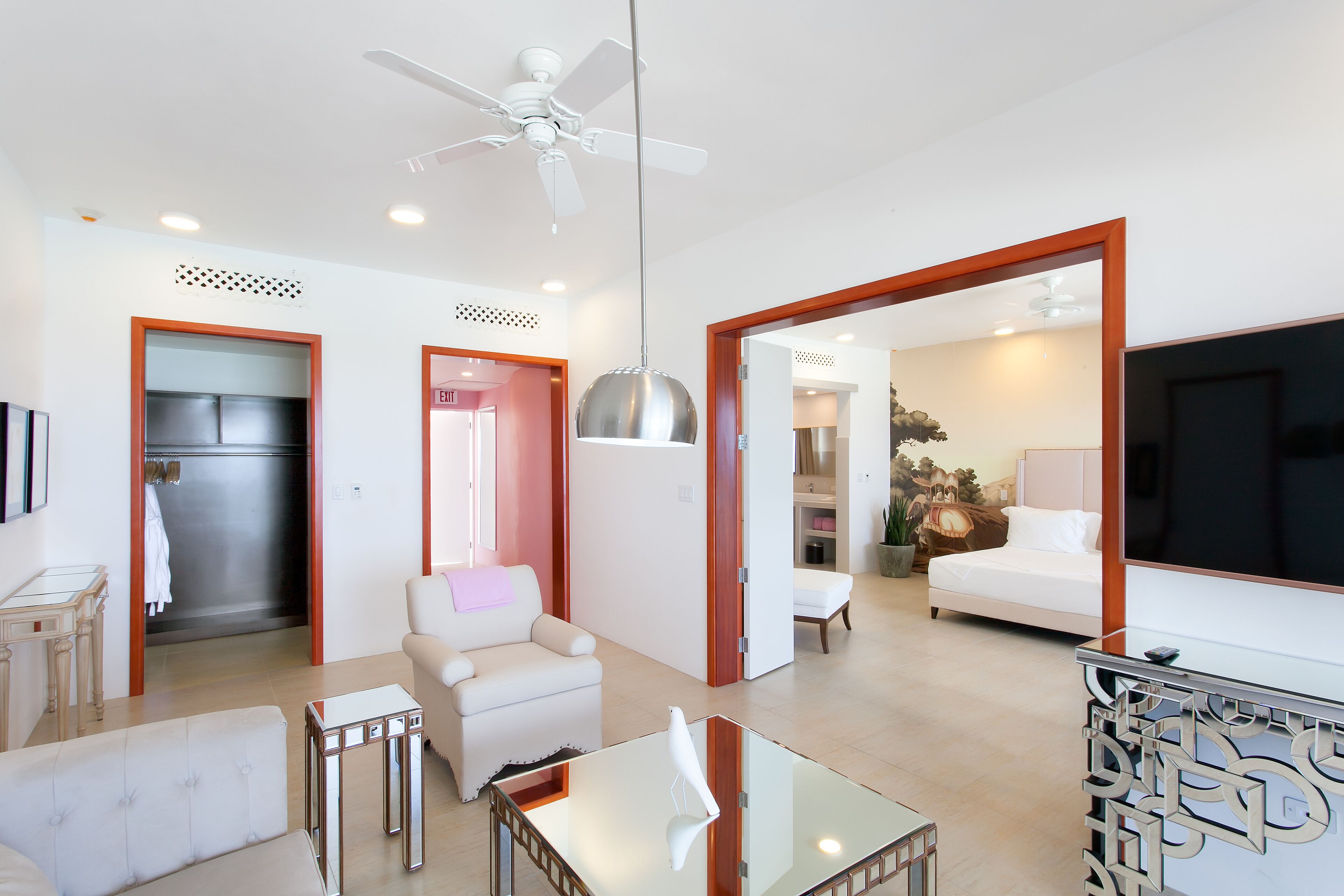 A spacious living area with a sofa, armchair, and a glass coffee table. There is a ceiling fan, a TV, and a bed in the adjacent room.