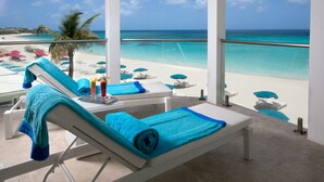 Un balcón con vista a una playa y al océano. Hay sillones de playa con toallas azules, una mesa con bebidas y una palmera.