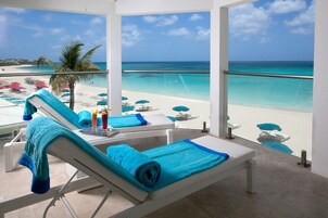 A balcony with a view of a beach and ocean. There are lounge chairs with blue towels, a table with drinks, and a palm tree.