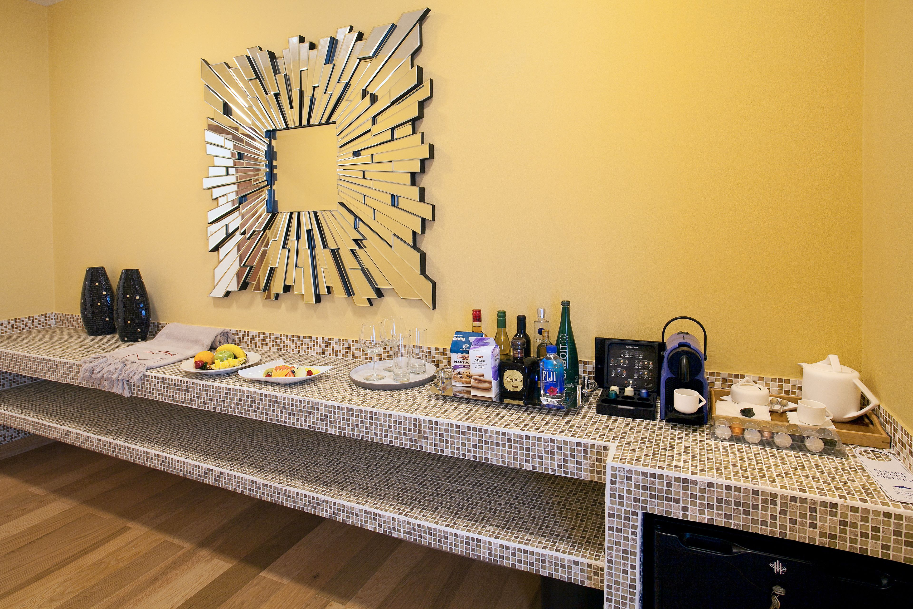 Zona de recepción de hotel con una decoración moderna, que incluye un espejo montado en la pared y un mostrador con diversos servicios como una máquina de café, botellas y vasos.