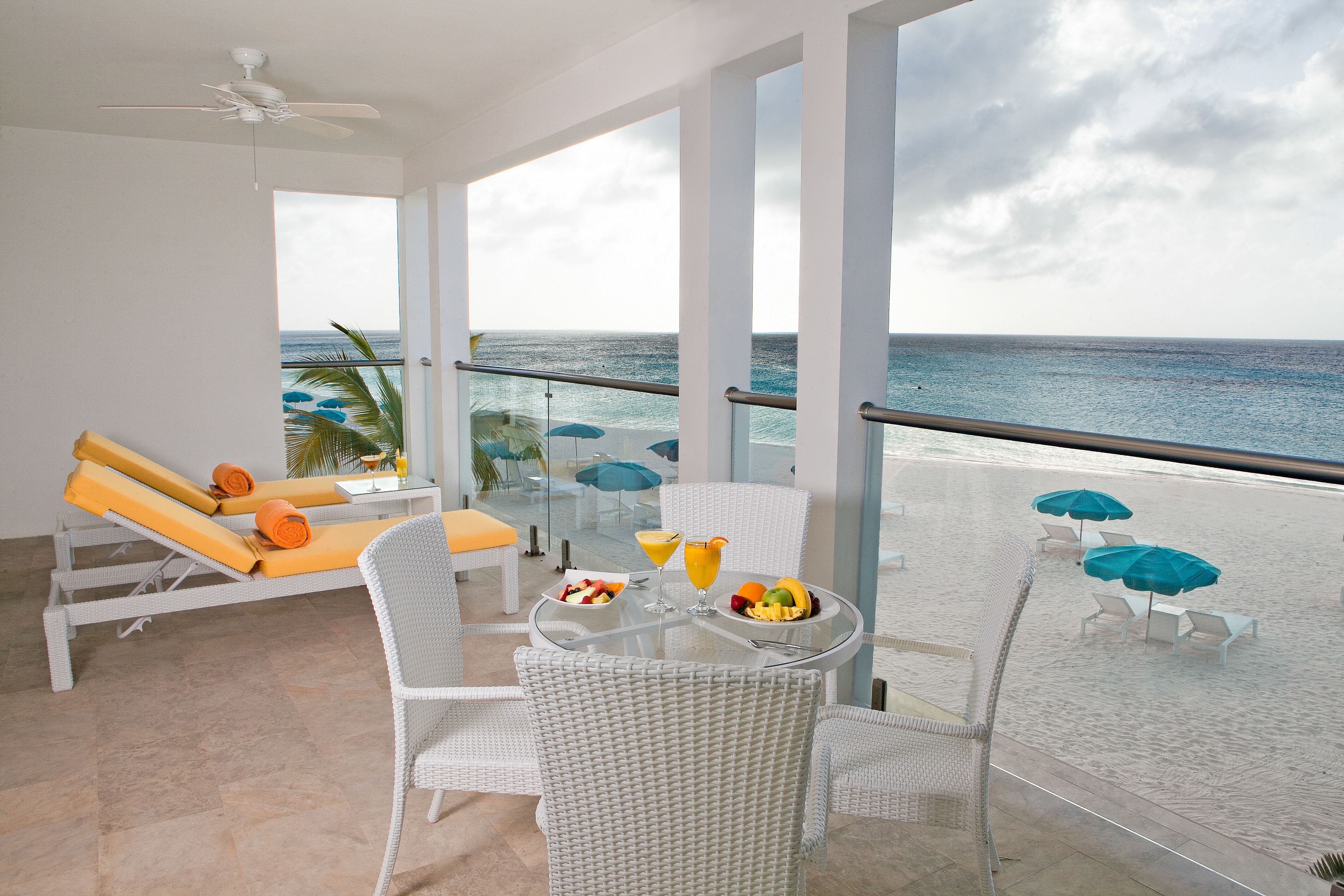 Varanda com mobiliário de vime branco, uma mesa de vidro com frutas e bebidas, e vista para o oceano.